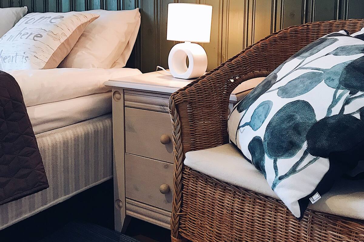A cozy bedroom with a bed, a nightstand, a wicker chair, and a lamp.