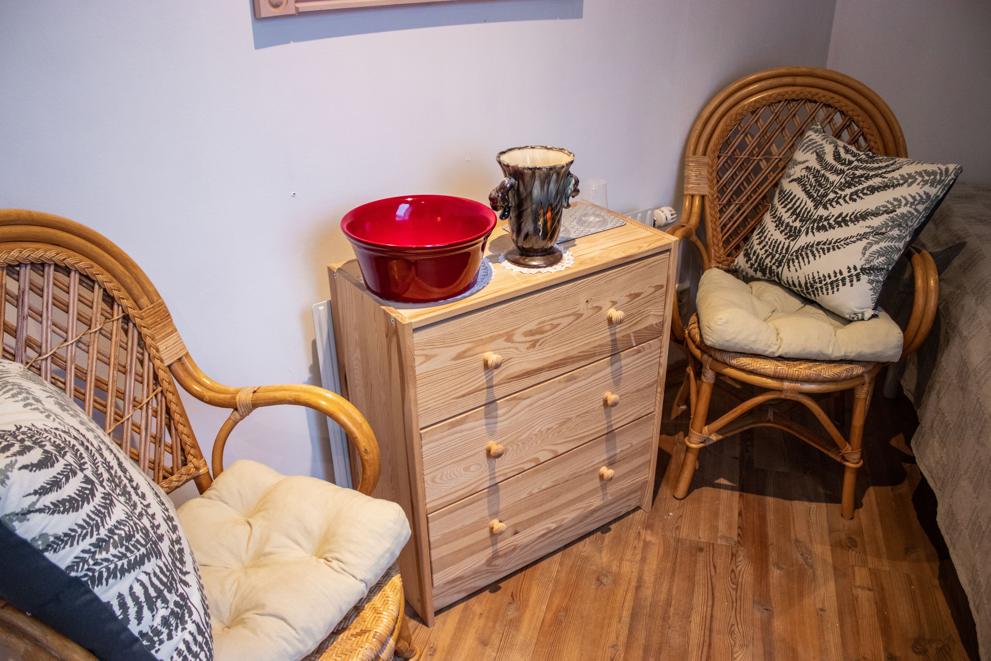 Une pièce comprenant une commode en bois, une chaise en osier avec un coussin et un bol rouge posé sur le dessus.