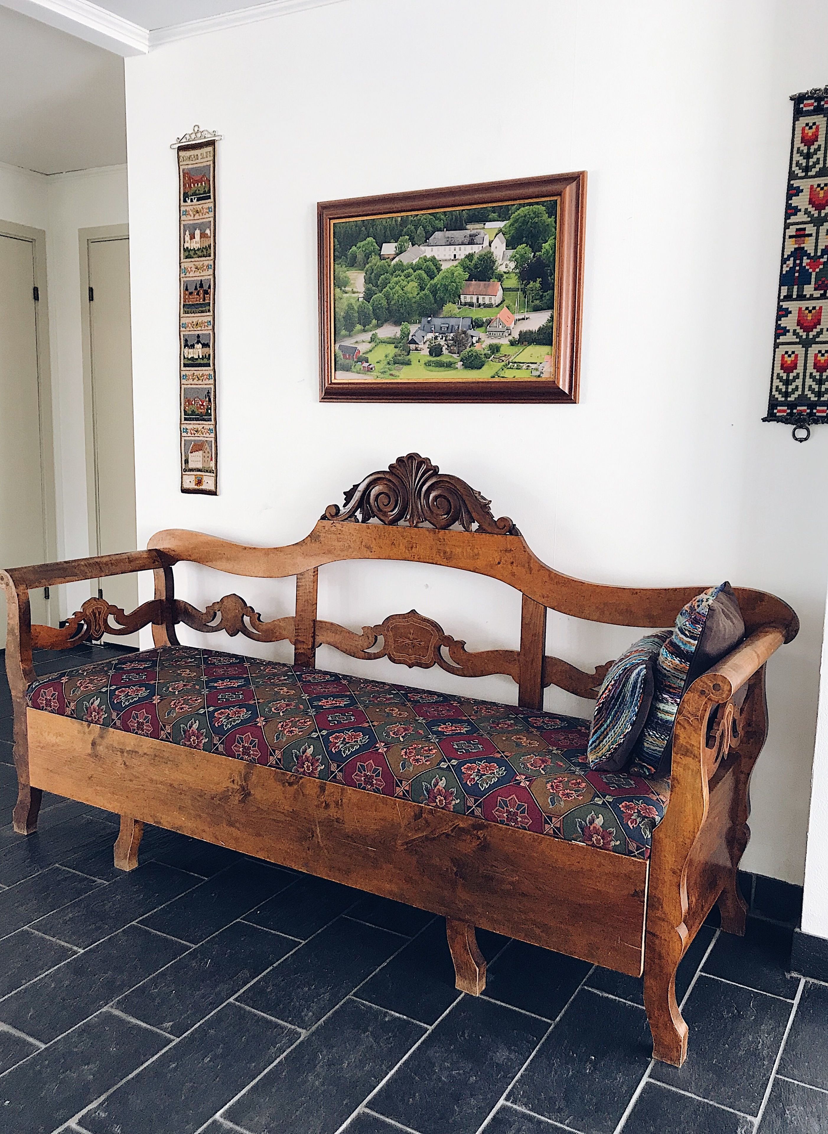 A wooden bench with a patterned cushion in a room with a tiled floor, a framed landscape painting on the wall, and a decorative wall hanging.