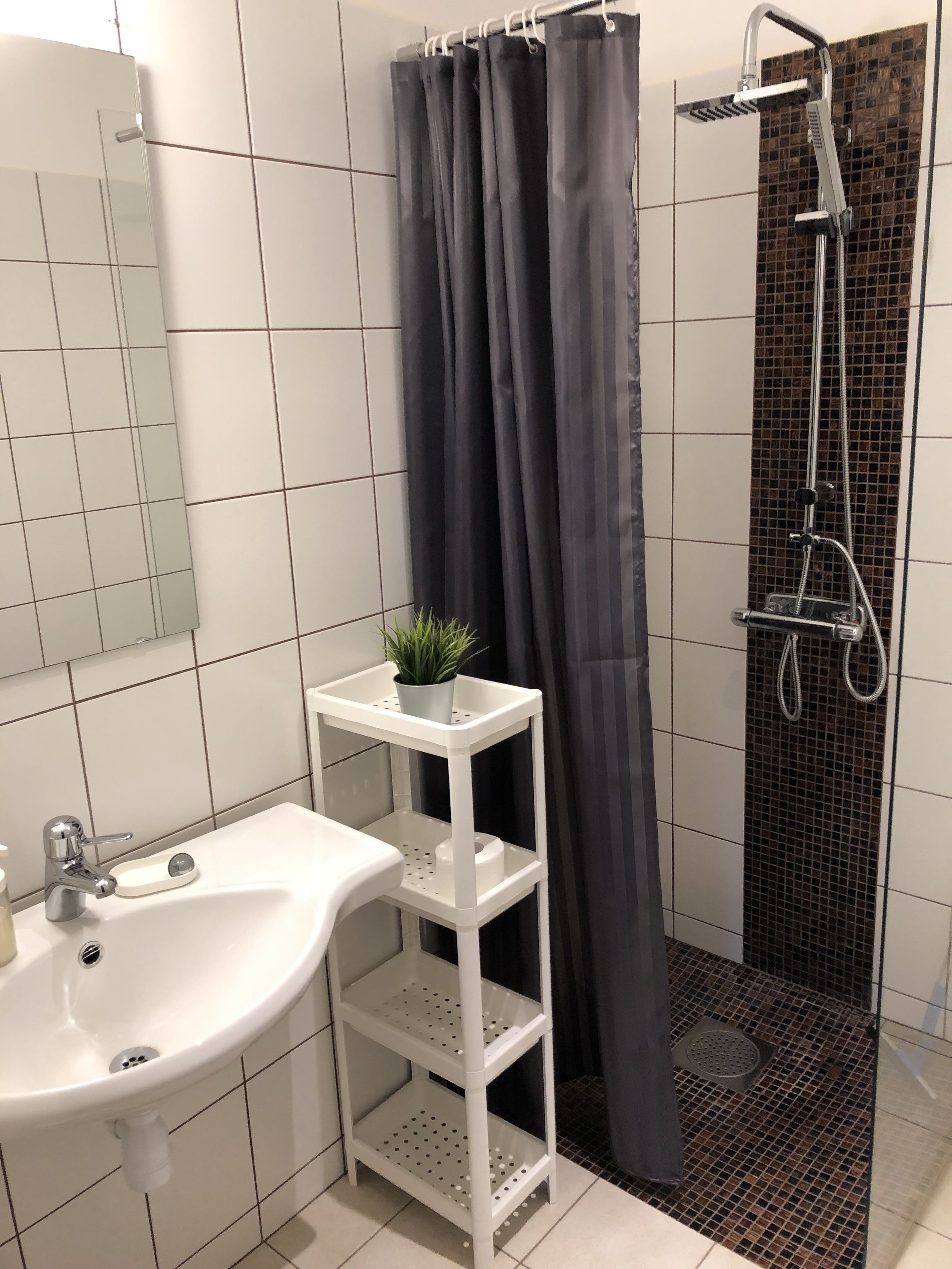 Un baño con un lavamanos blanco, un espejo, una ducha con cortina negra y una planta pequeña en un estante.