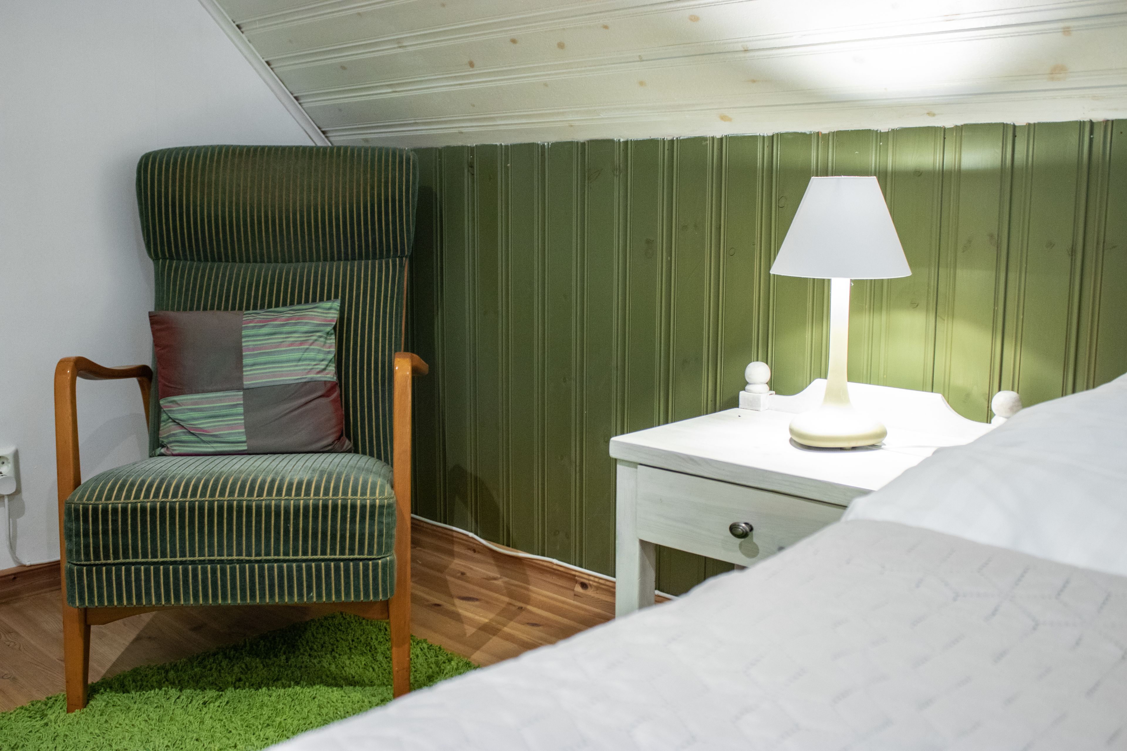 Una habitación con una silla verde a rayas, una mesita de noche blanca con una lámpara y una cama con ropa de cama blanca.