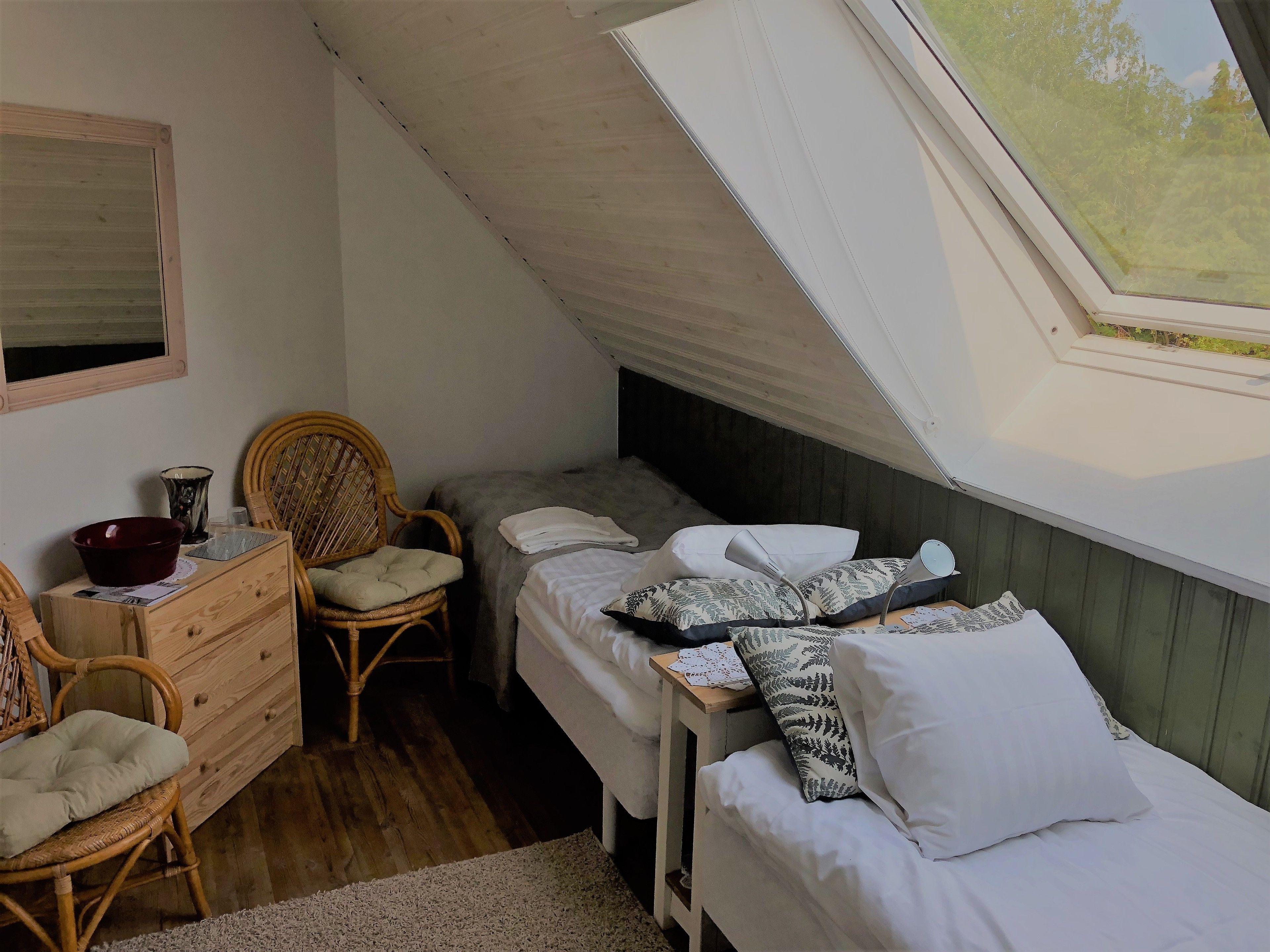 A small, cozy bedroom with a single bed, a wooden dresser, a wicker chair, and a skylight.