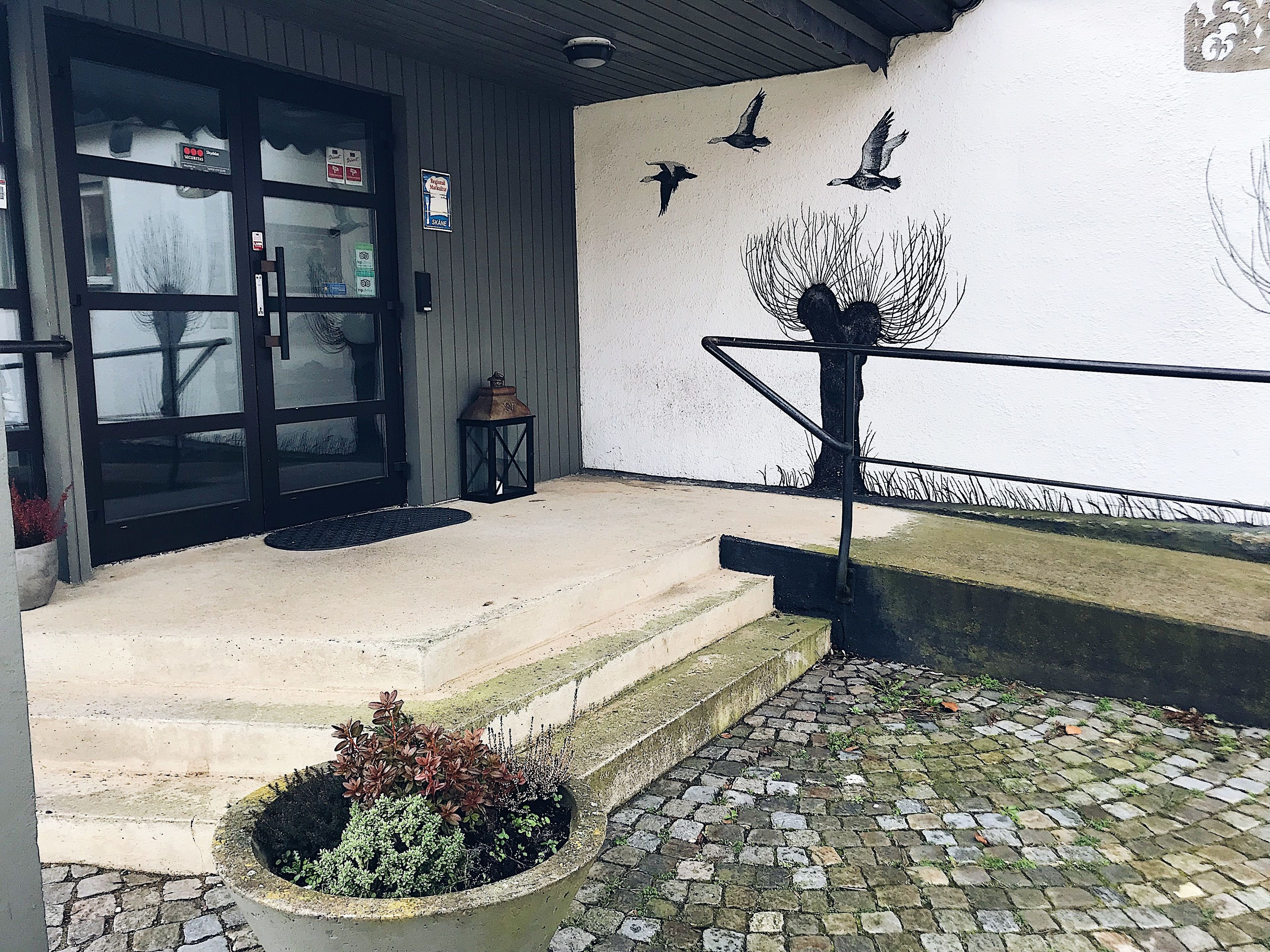 Entrada de un edificio con puerta negra, un pequeño espacio de asientos al aire libre, un farol y una escultura de pájaro en la pared.