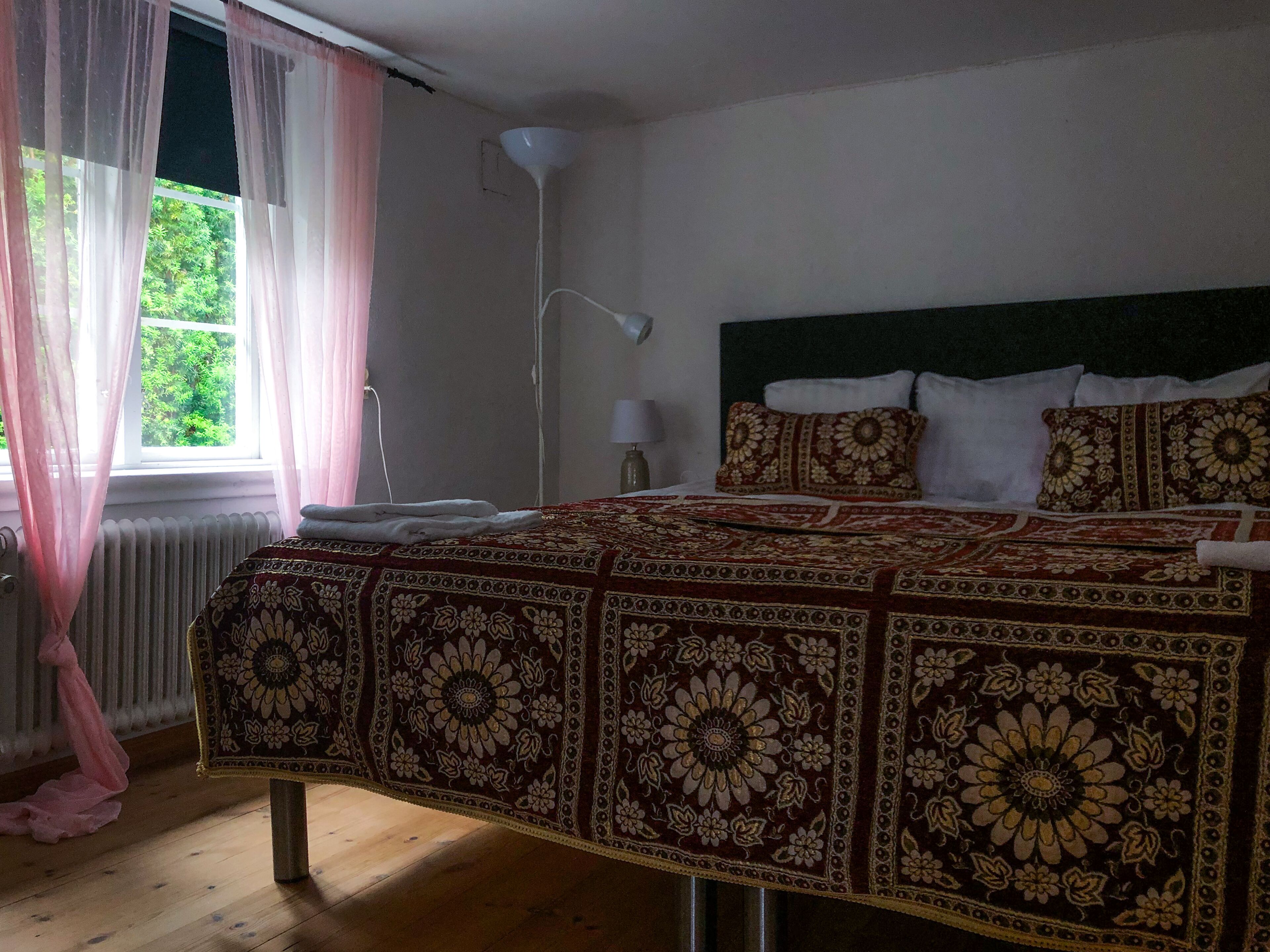 A bedroom with a large bed, a window with sheer curtains, a radiator, and a floor lamp.