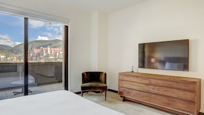 Une chambre d’hôtel avec un grand lit, une commode en bois, une chaise, un téléviseur fixé au mur et une porte-fenêtre coulissante donnant sur un balcon avec vue sur la ville.