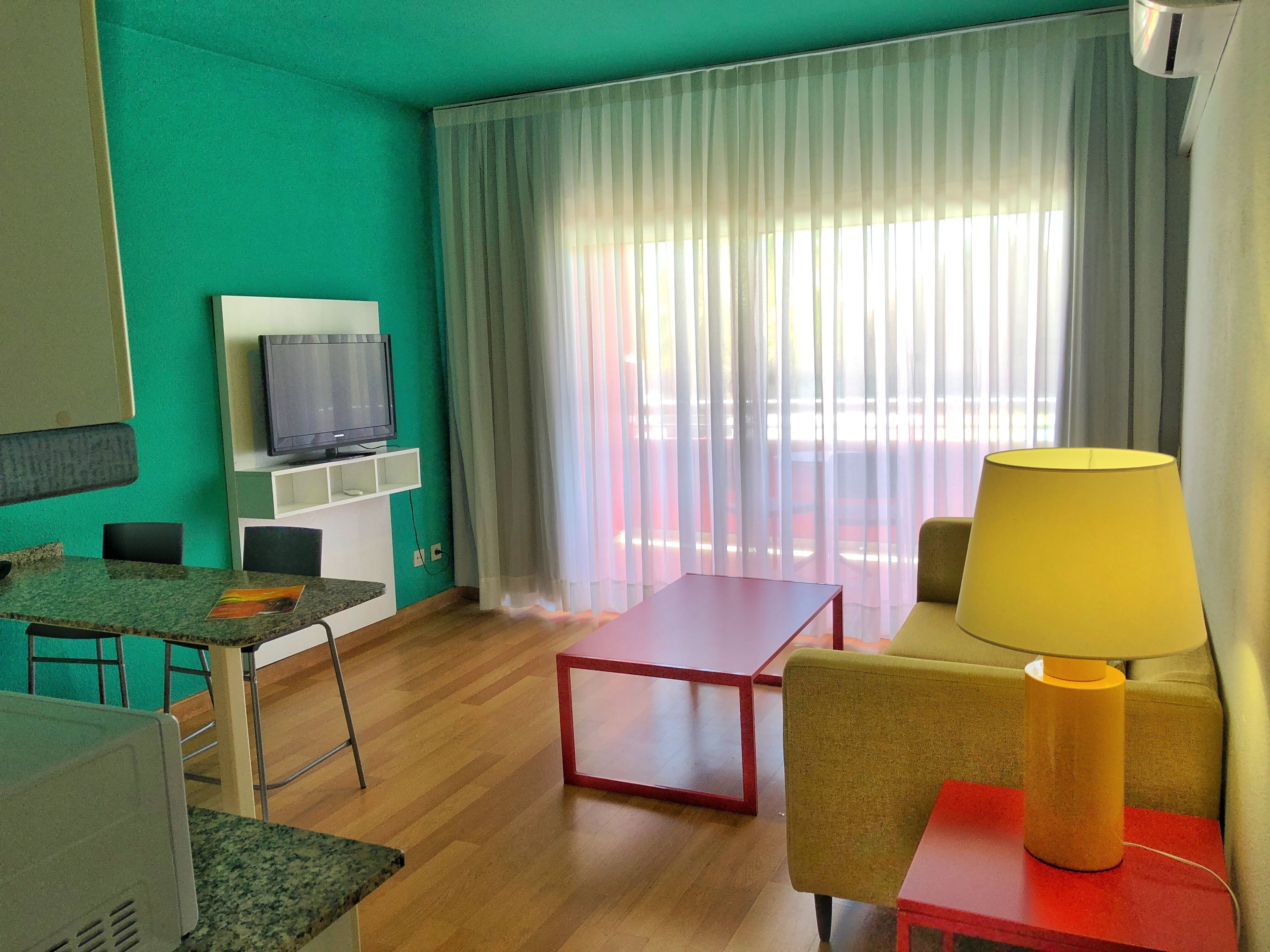 A room with a television on a wall, a dining table with chairs, a sofa with a lamp, and a large window with curtains.