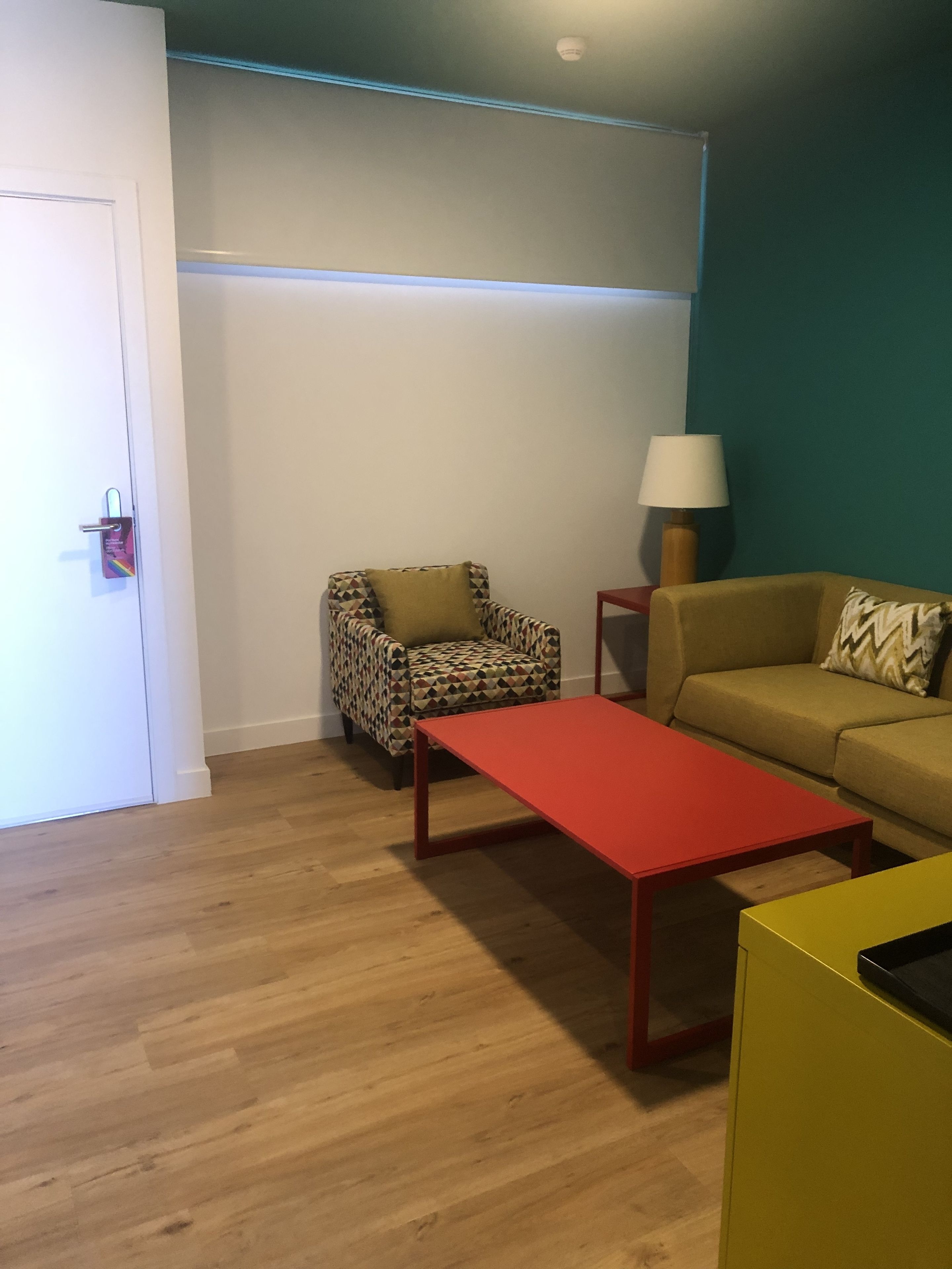 Une pièce avec un plancher en bois, une table rouge, un fauteuil à motifs, un canapé beige et une lampe.