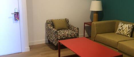 Une pièce avec un plancher en bois, une table rouge, un fauteuil à motifs, un canapé beige et une lampe.