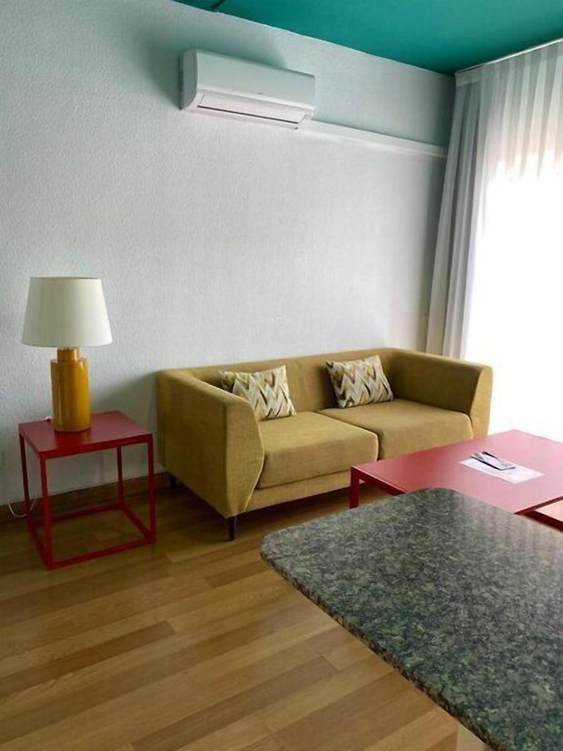A living room with a sofa, a small table with a lamp, a red table, and a patterned rug.