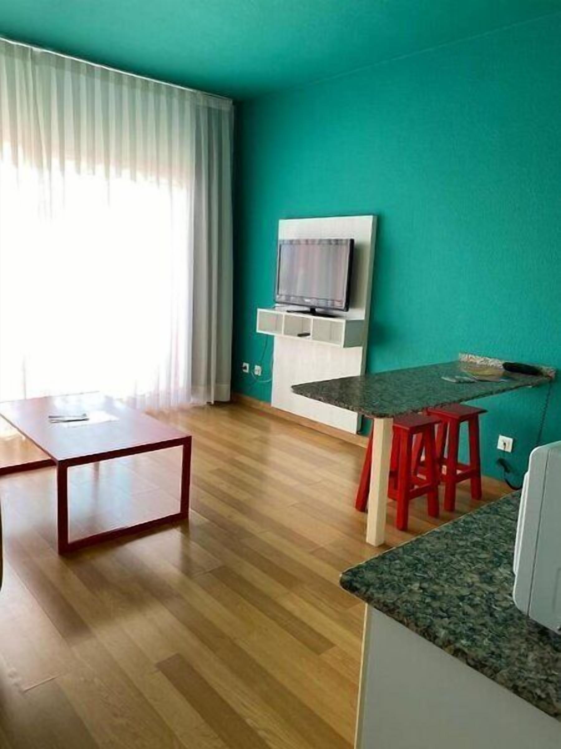 A compact living space with a wooden floor, a dining table with red stools, a TV mounted on a white wall, and a window with white curtains.