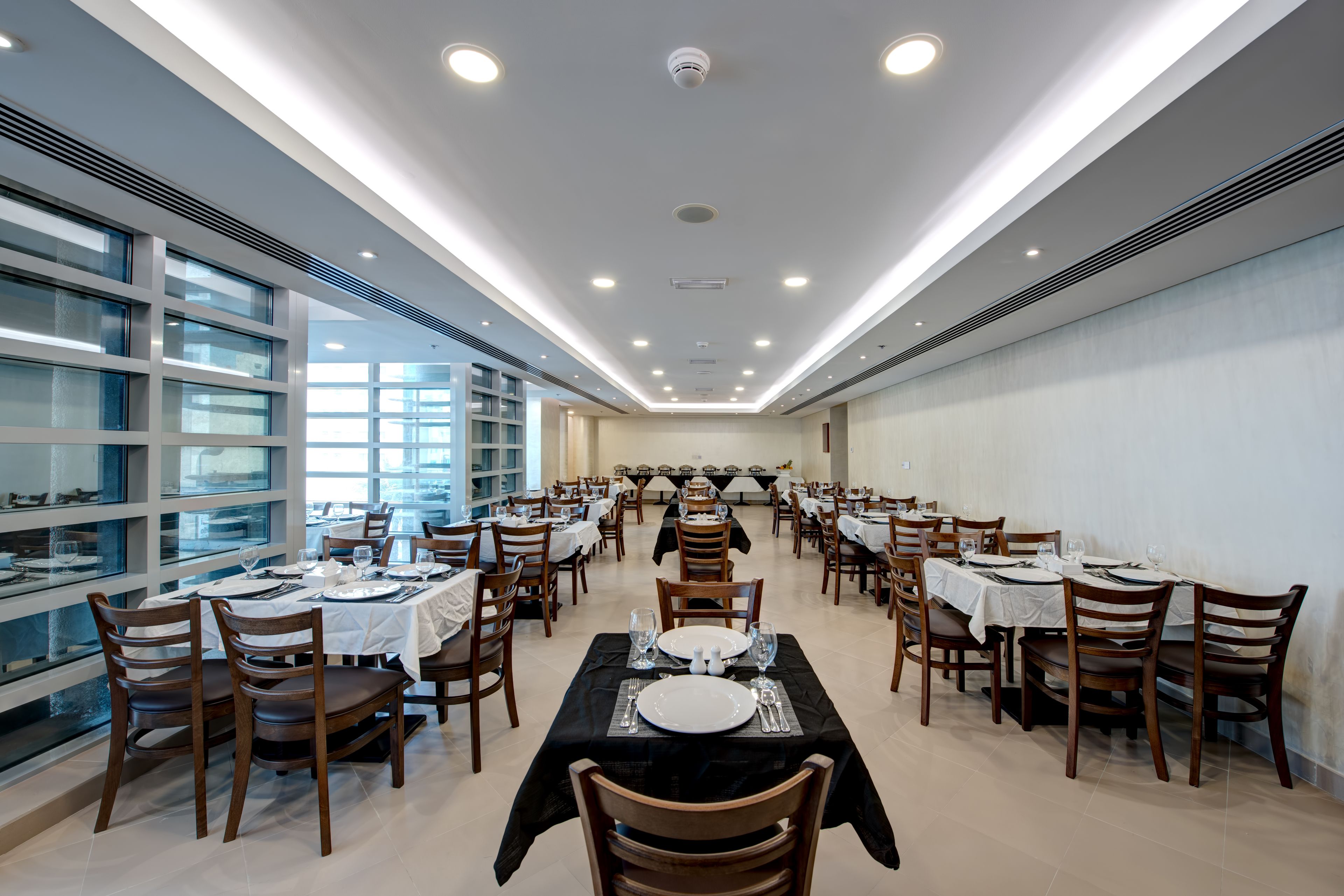 A spacious dining area with multiple tables and chairs, large windows on one side, and a modern ceiling design.