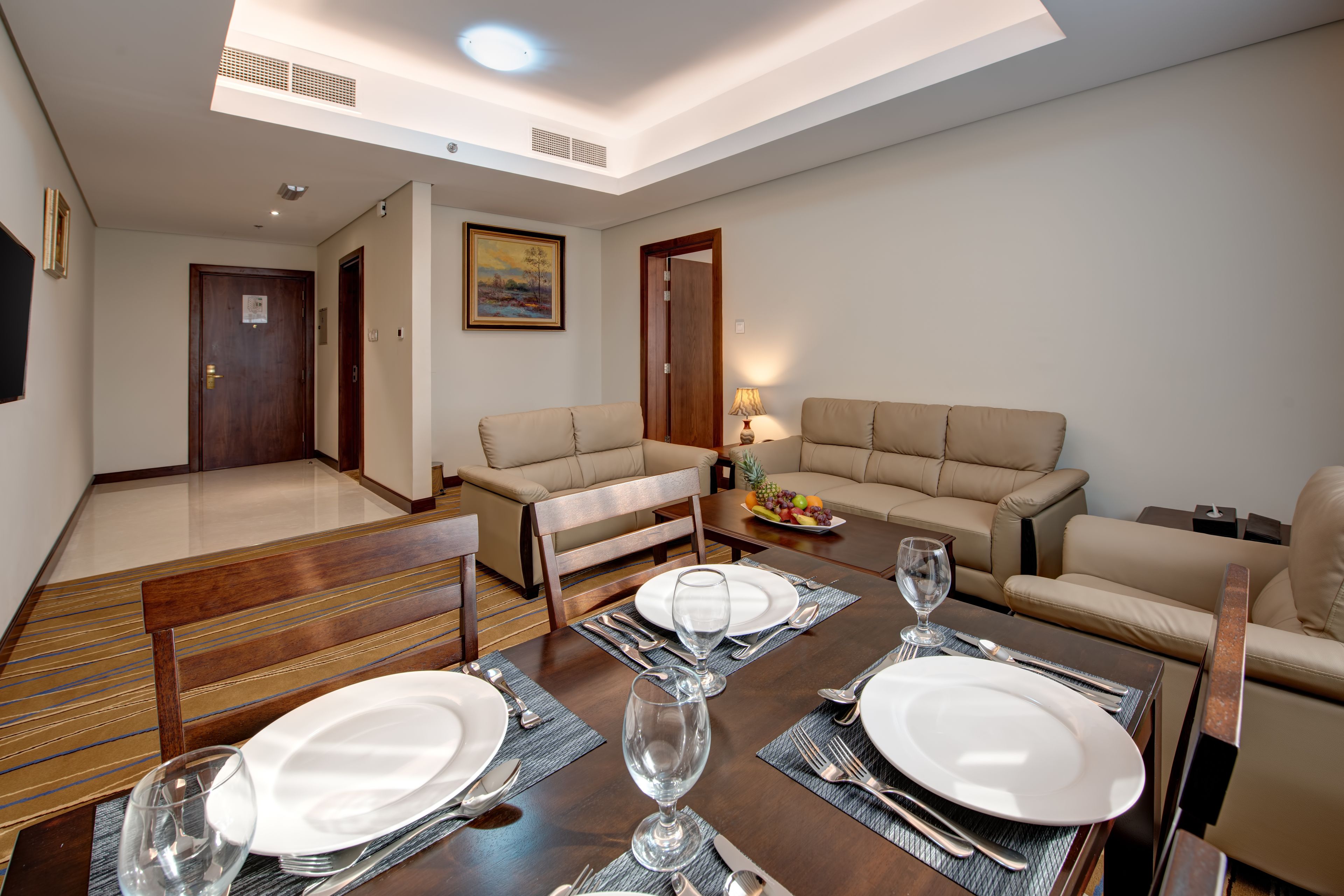 A dining area with a wooden table set for four, including plates, cutlery, and glasses. There are two sofas and a small side table with a fruit bowl.