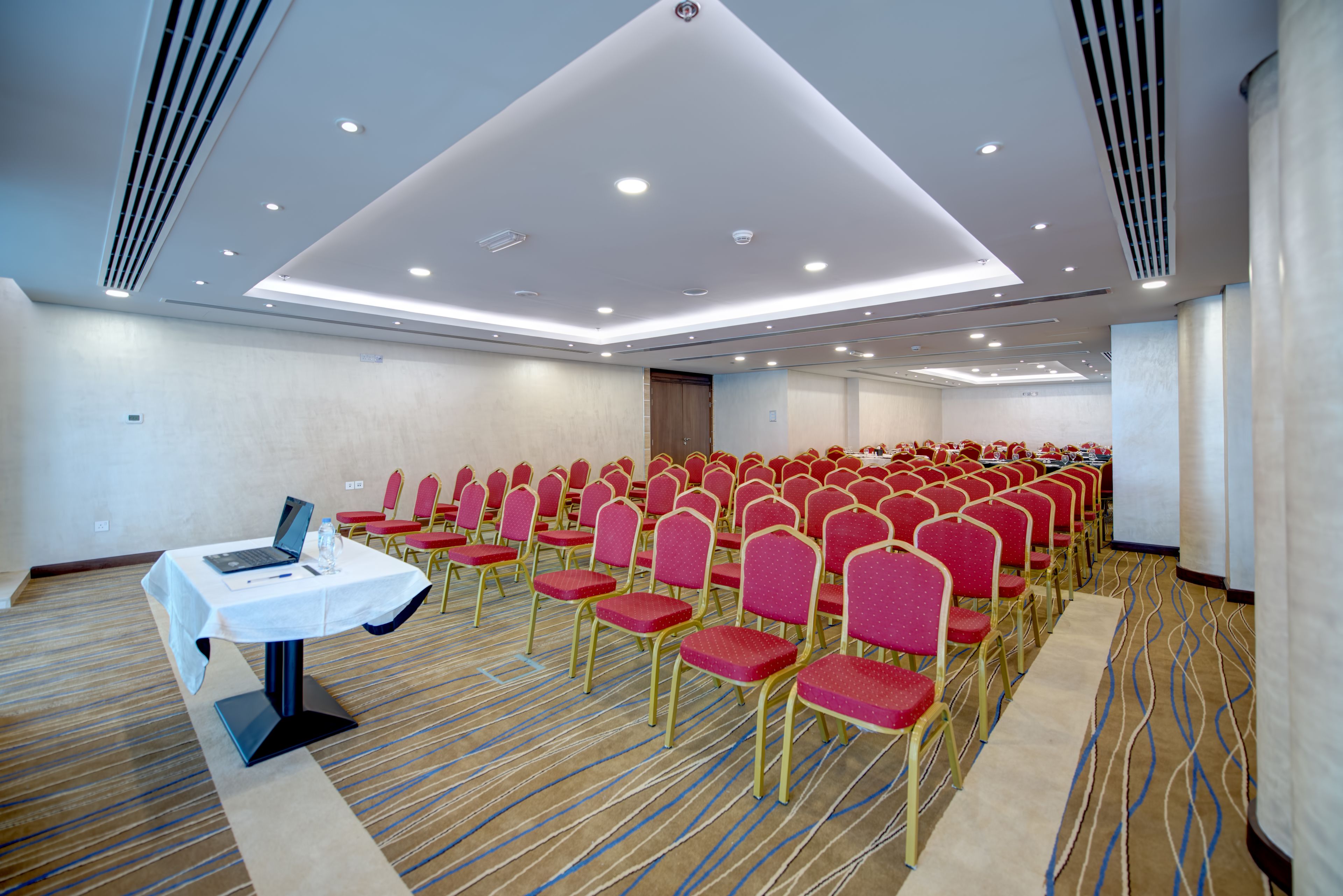 Une salle de conférence avec des rangées de chaises rouges, une table blanche sur laquelle se trouve un ordinateur portable, et un sol recouvert de moquette.
