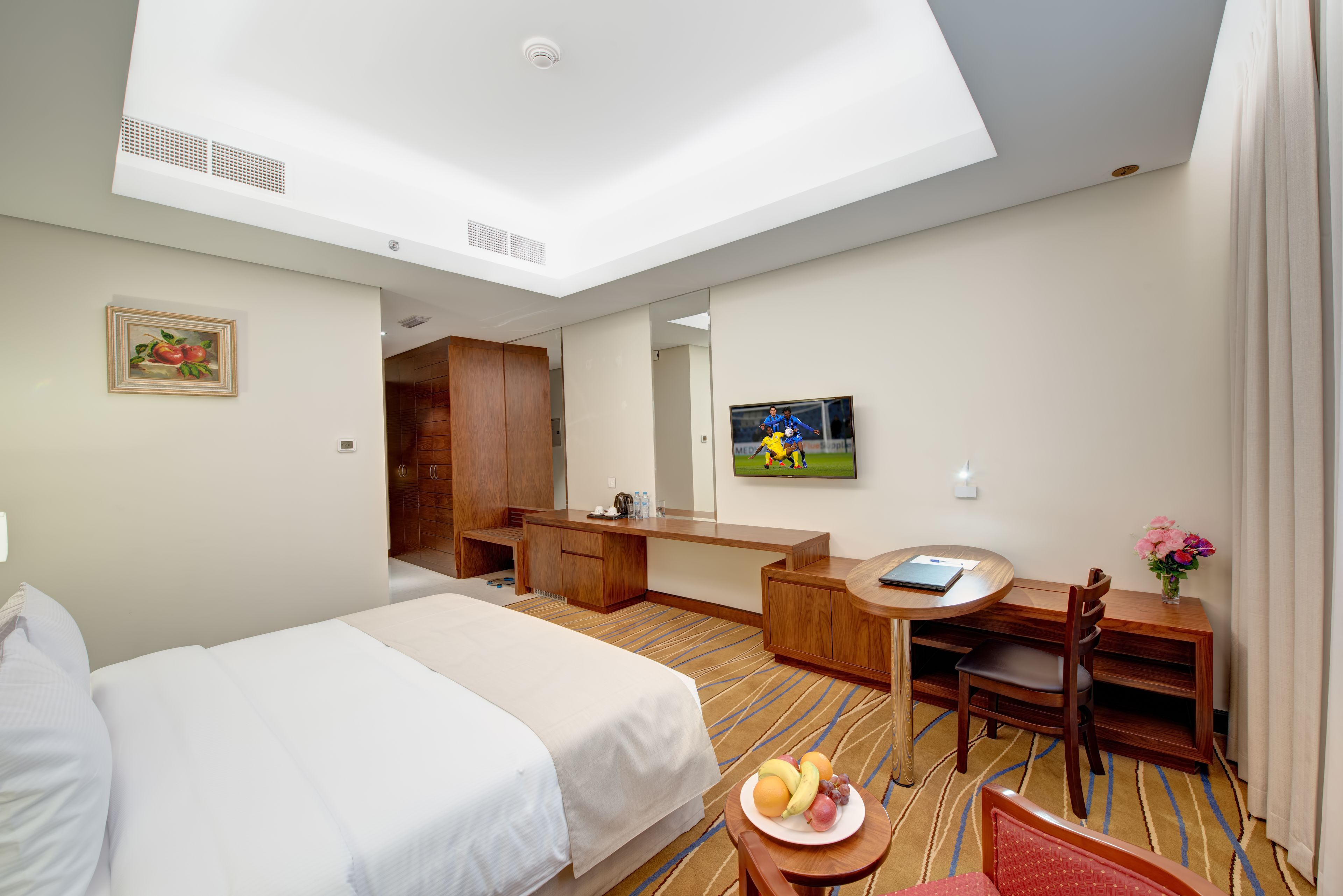 A hotel room with a bed, a desk, a chair, a small table with fruit, and a TV on the wall.