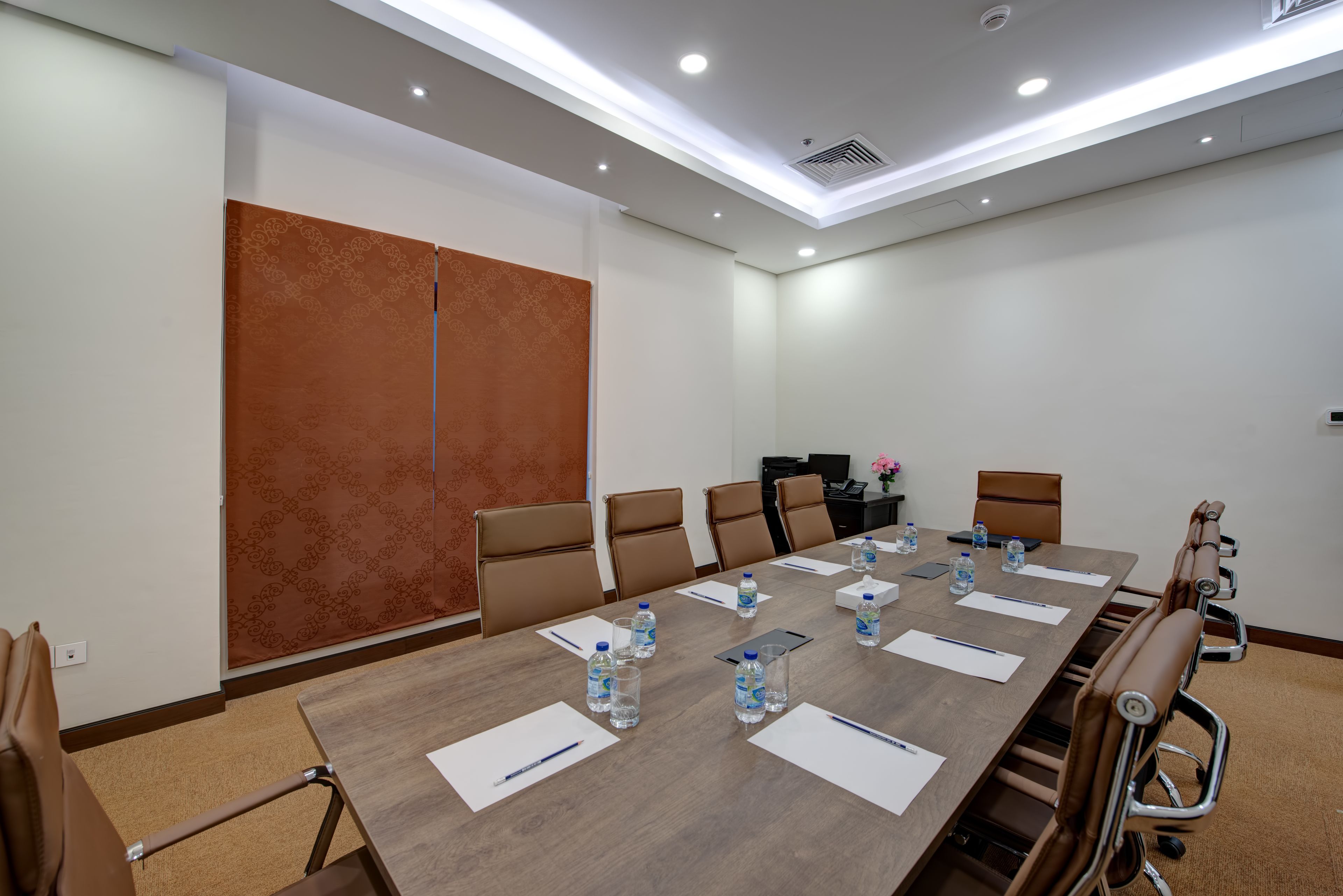 Una sala de conferencias con una larga mesa, sillas de cuero marrón, botellas de agua y una computadora portátil.