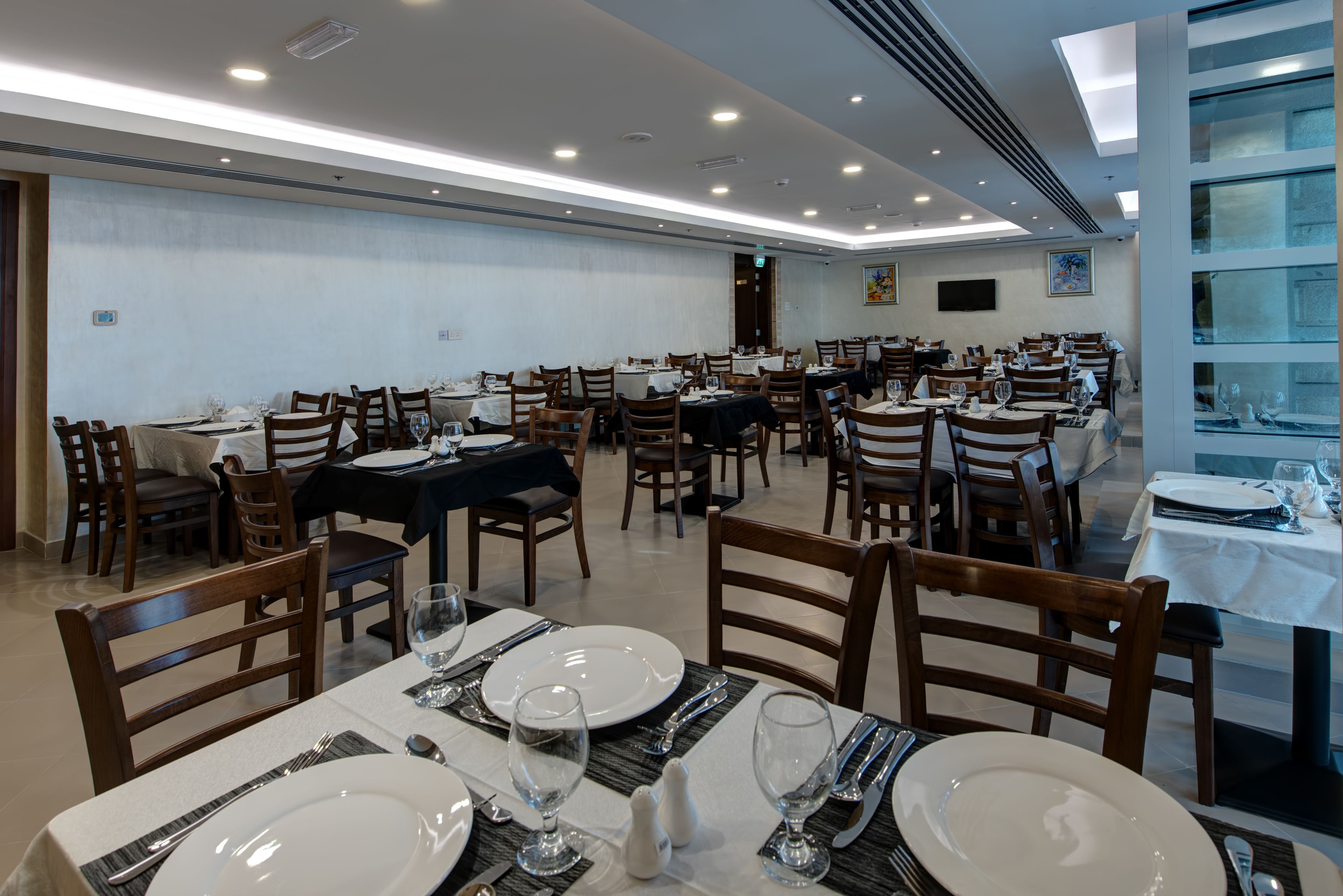 Uma sala de jantar espaçosa com várias mesas e cadeiras, bem iluminada por luzes de teto, e uma grande janela que oferece luz natural.