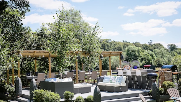 Un patio exterior con una pérgola de madera, asientos acolchados y una parrilla.