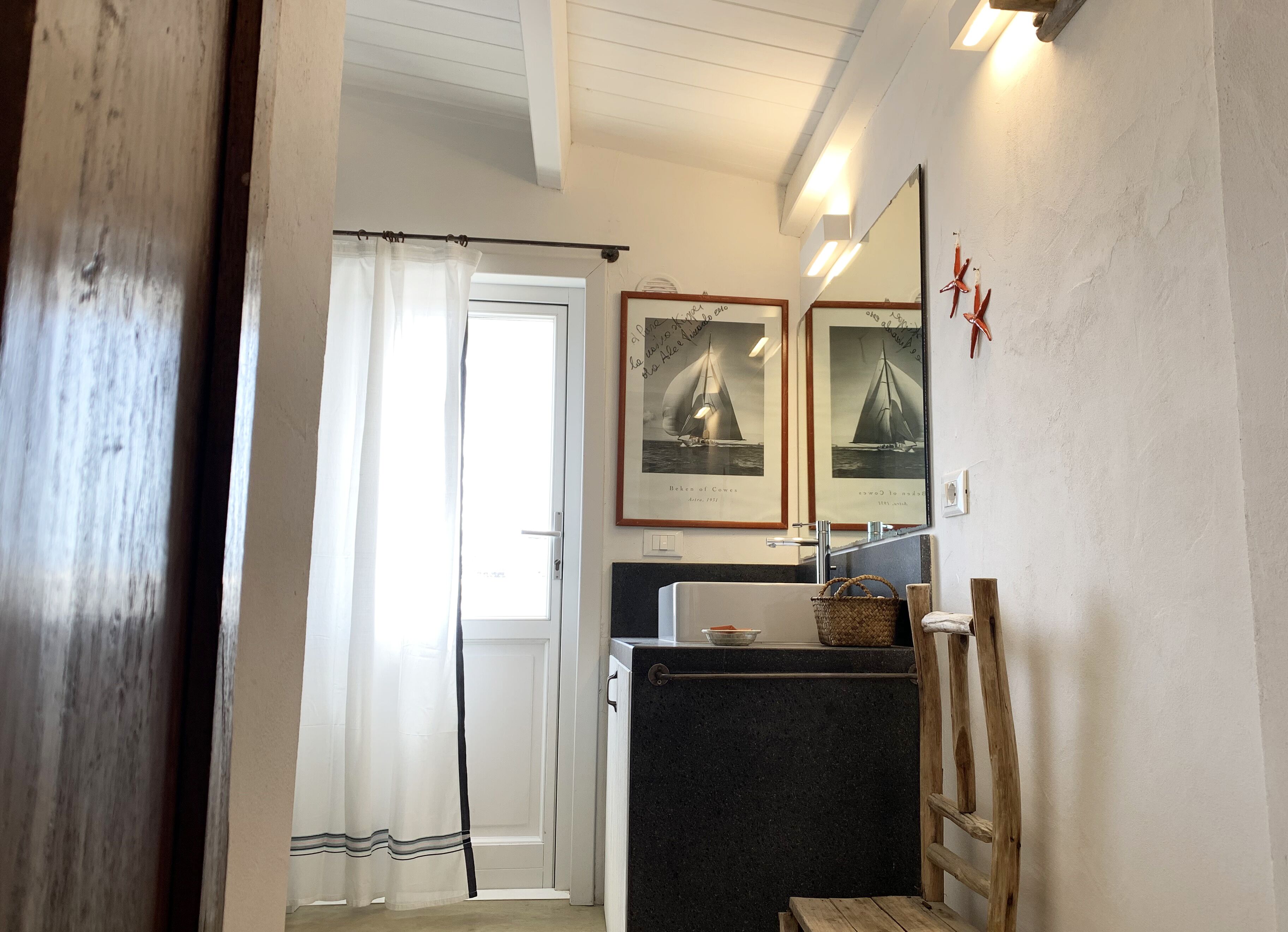 Um banheiro com porta branca, cortina de chuveiro, pia com espelho e quadros nas paredes.