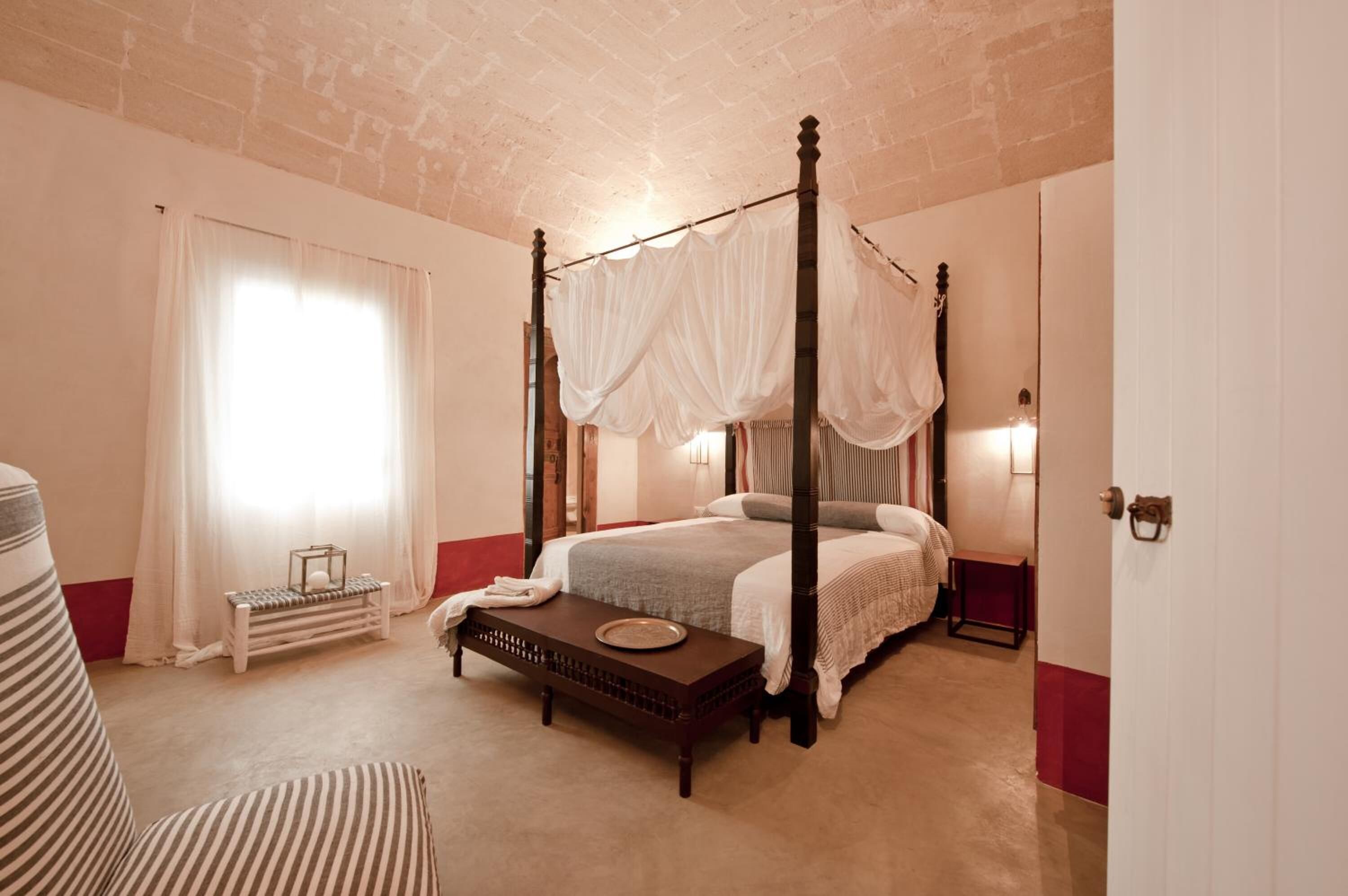 A four-poster canopy bed with a bedside table and bench, a window with curtains, and a brick ceiling.