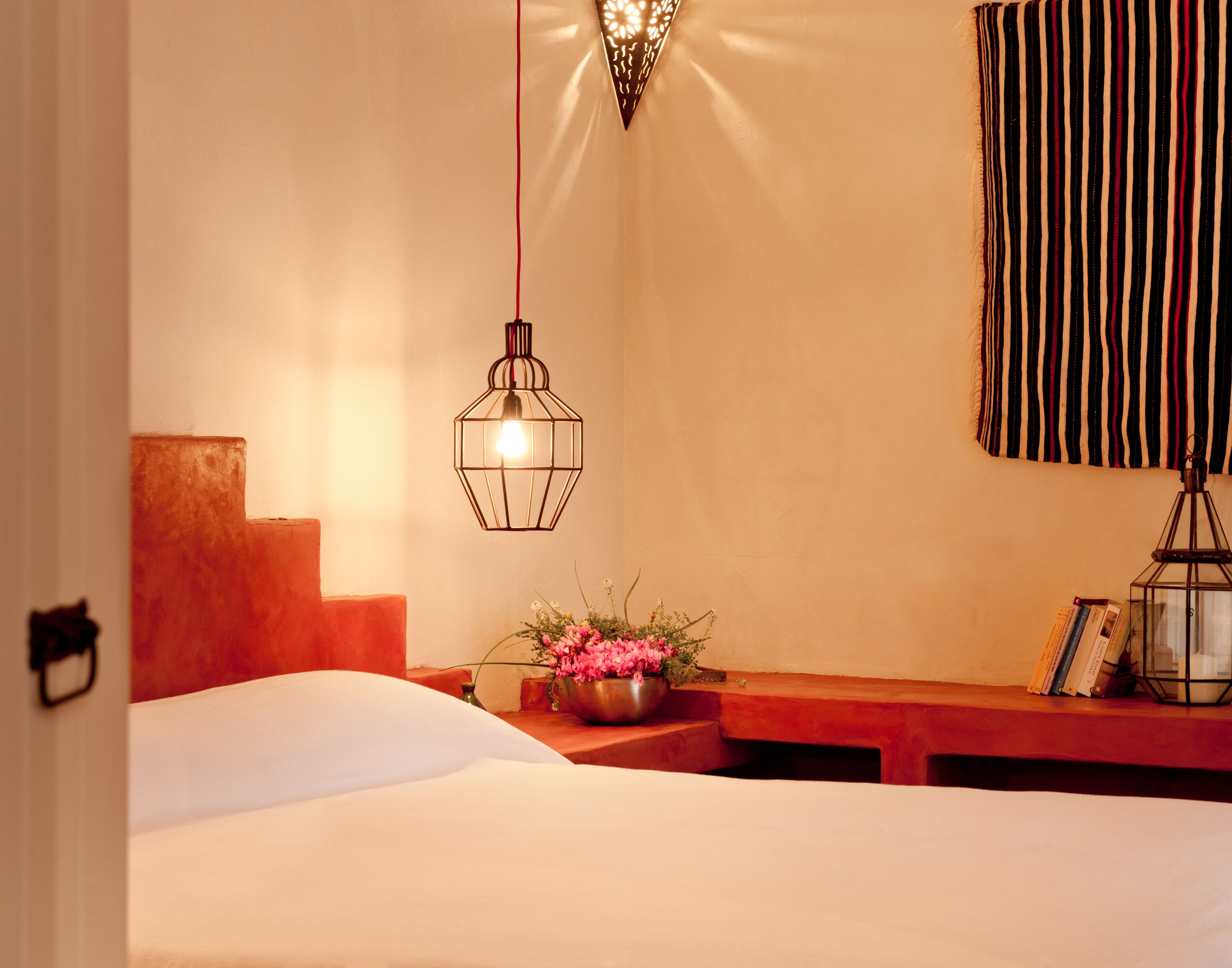 A bedroom with a bed, a nightstand with a lamp, a window with striped curtains, and a wall-mounted light fixture.