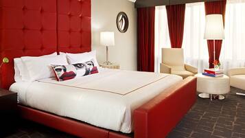 Una habitación de hotel con una cama grande, cabecera tapizada en rojo, una mesita de noche redonda con lámpara, una silla y una pequeña mesa con libros y un jarrón.