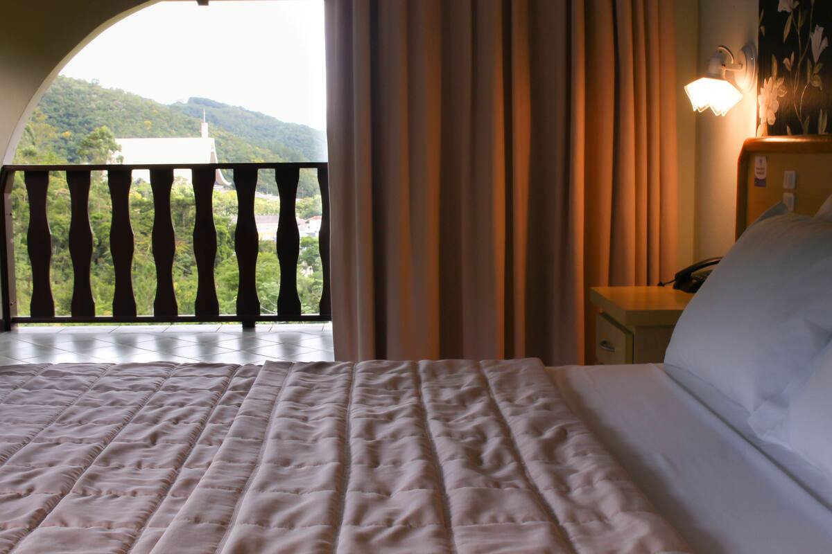 A hotel room with a bed, a nightstand, a lamp, and a view of a balcony with a railing.