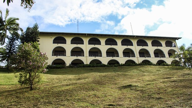 Un edificio con varias ventanas arqueadas y techo plano, rodeado de césped y algunos árboles.