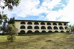 Un edificio con varias ventanas arqueadas y techo plano, rodeado de césped y algunos árboles.