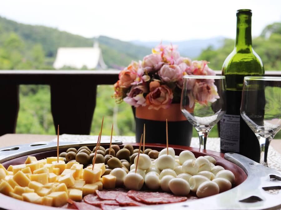 Uma tábua de queijos com diversos tipos de queijo e azeitonas, uma garrafa de vinho e duas taças sobre uma mesa com uma vista panorâmica ao fundo.