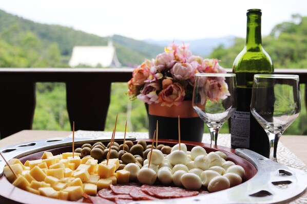 Una tabla de quesos con varios tipos de queso y aceitunas, una botella de vino y dos copas sobre una mesa con vistas panorámicas al fondo.