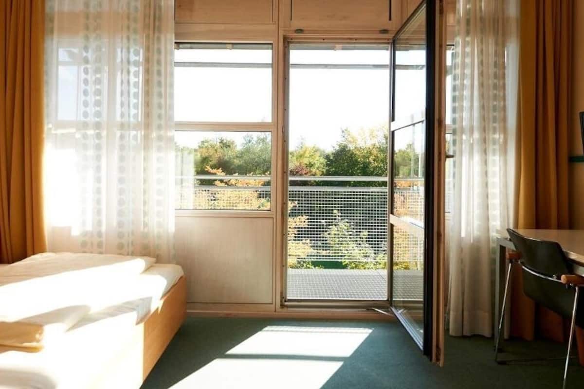 A bedroom with a bed, a desk, and a view of a balcony with a railing.