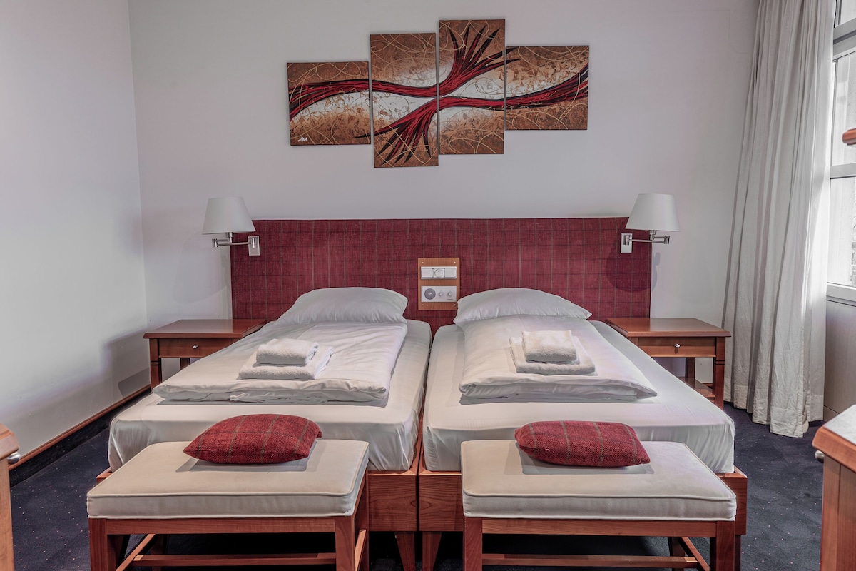 Een hotelkamer met twee eenpersoonsbedden, een rode hoofdbord, nachtlampjes, een kunstwerk aan de muur en een raam met gordijnen.