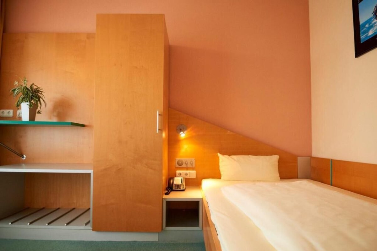 A hotel room with a bed, a nightstand, a shelf with a plant, and a wardrobe.