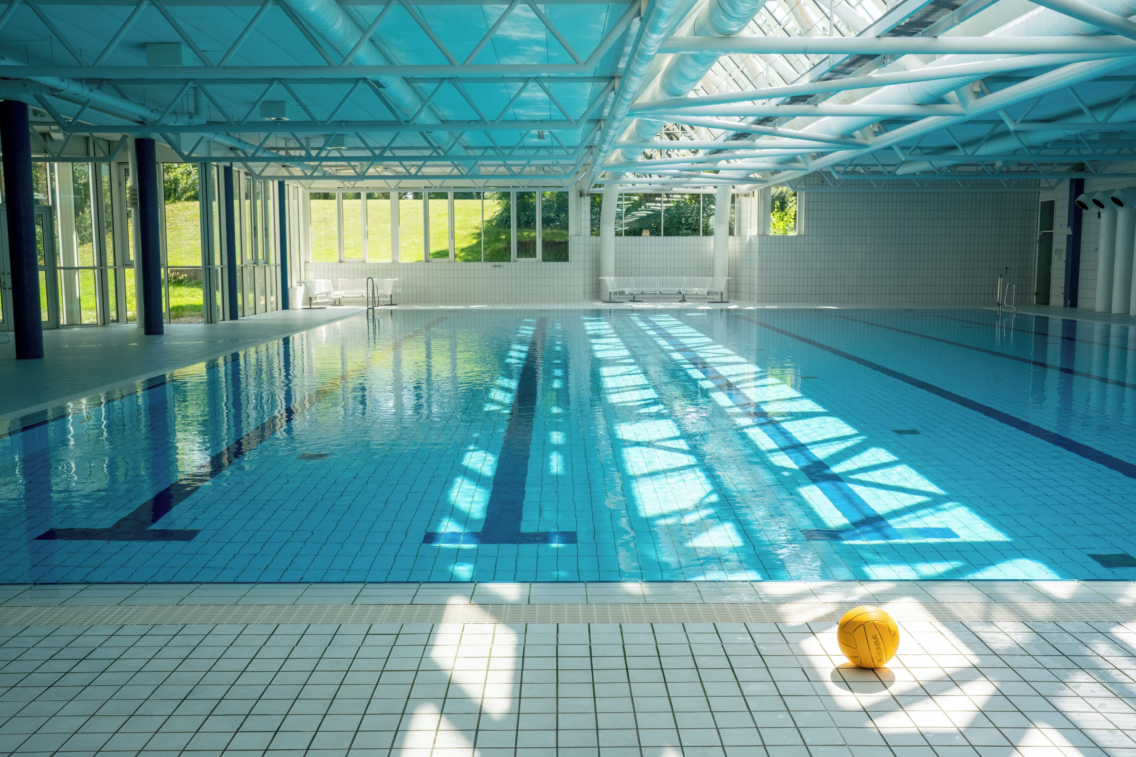 Una piscina interior con agua azul cristalina, suelo de baldosas y una pelota amarilla en el borde.