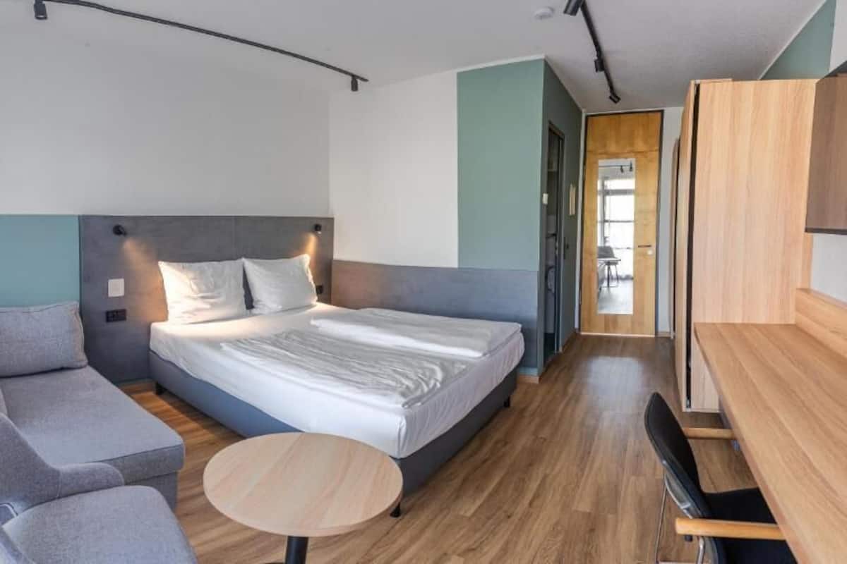 A modern hotel room with a bed, a sofa, a small round table, and a wooden desk.