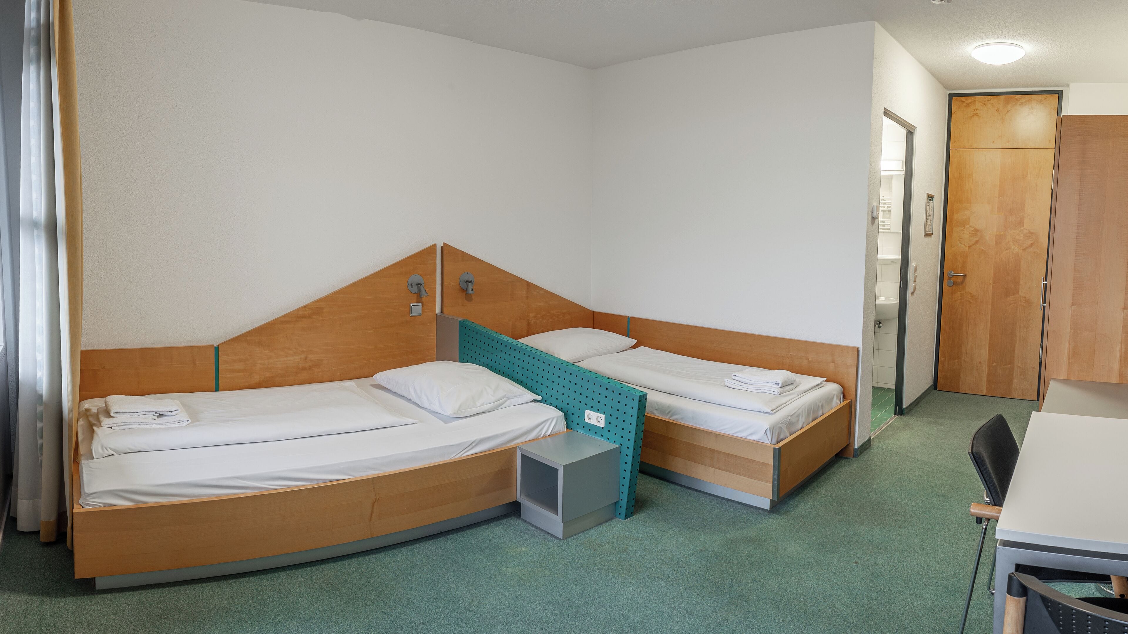 A compact hotel room with two wooden beds, a small desk, and a green carpet.