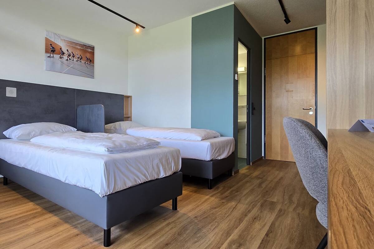 A modern hotel room with two single beds, a wooden desk, and a chair.