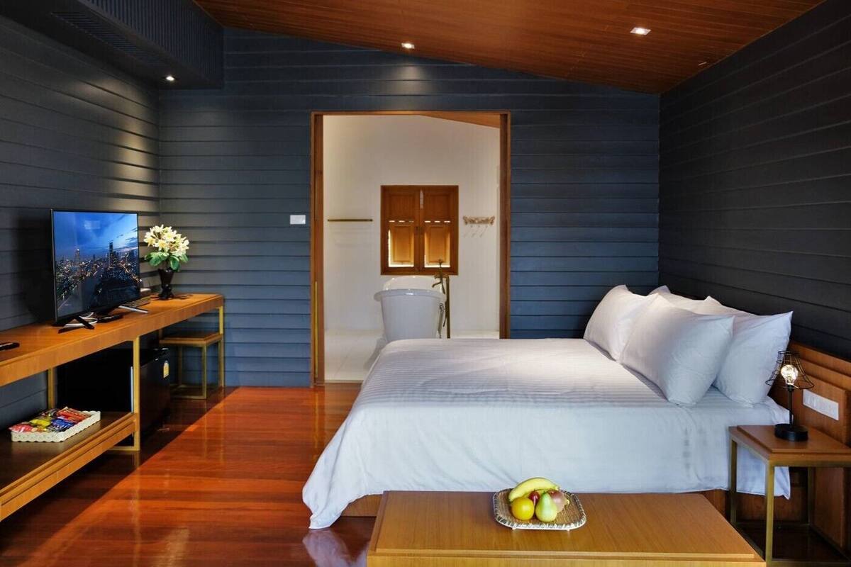 Une chambre moderne avec un plafond en bois, un grand lit avec une literie blanche, une table de chevet avec une lampe et un panier de fruits posé sur le sol.