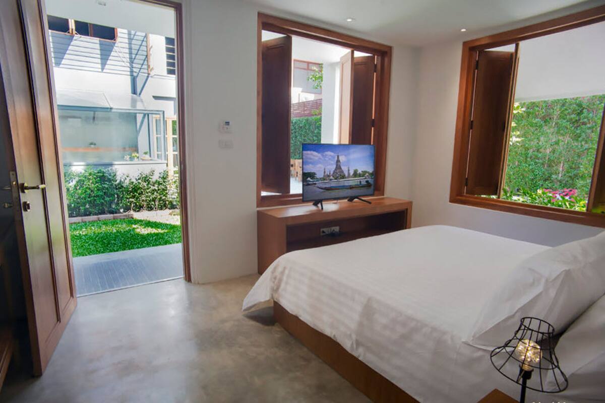 Une chambre à coucher avec un lit, un téléviseur posé sur un meuble en bois, une fenêtre avec une porte ouverte et une vue sur un jardin.