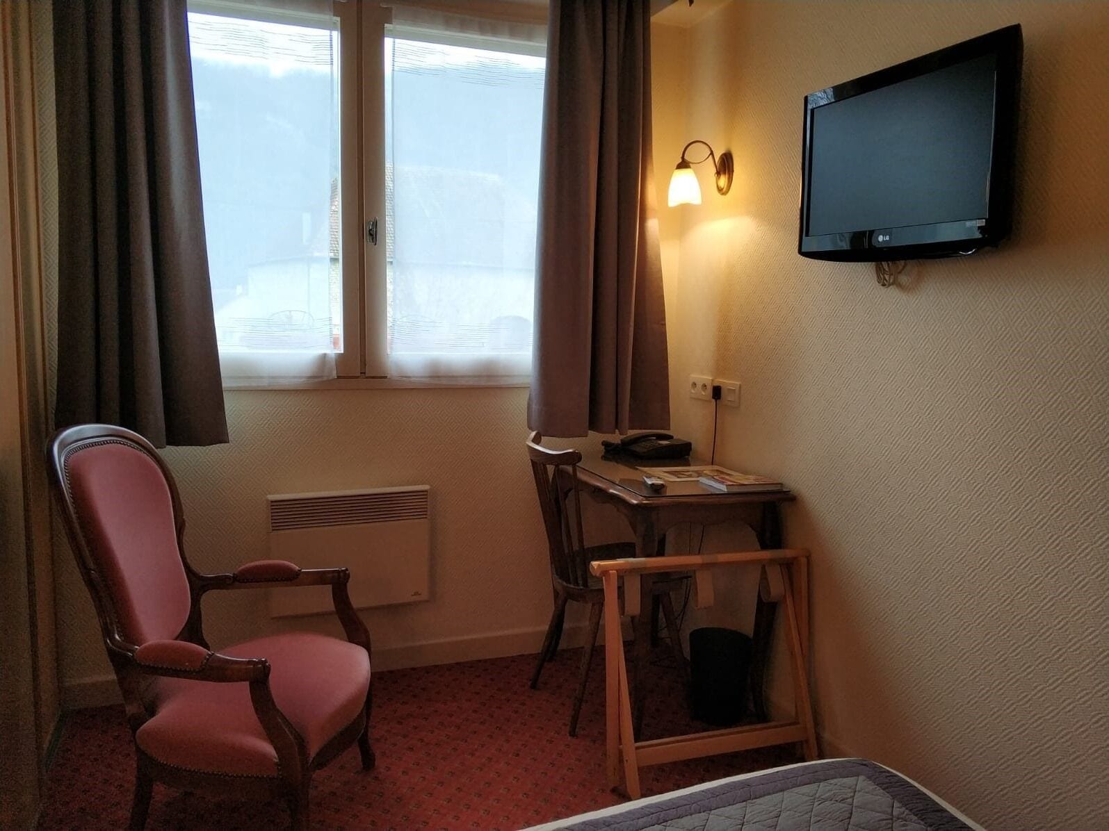 Une chambre d’hôtel avec un lit simple, une chaise en bois, une petite table avec un téléphone et un magazine, un téléviseur fixé au mur et une fenêtre avec des rideaux.