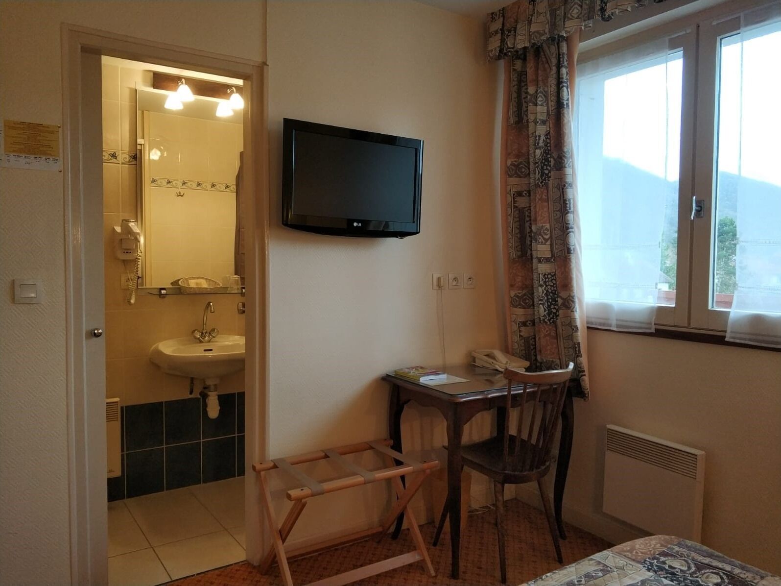 Ein kleines Zimmer mit Bett, Schreibtisch, Stuhl, Fernseher und einem Fenster mit Vorhängen. Ein Badezimmer ist durch eine offene Tür zu sehen.