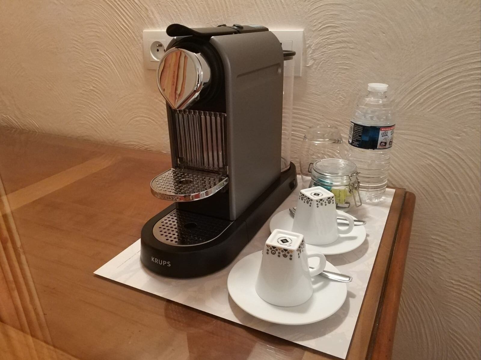 Une machine à café Krups est posée sur une table, à côté d’une bouteille d’eau, d’un bocal en verre et de deux tasses blanches avec couvercles.