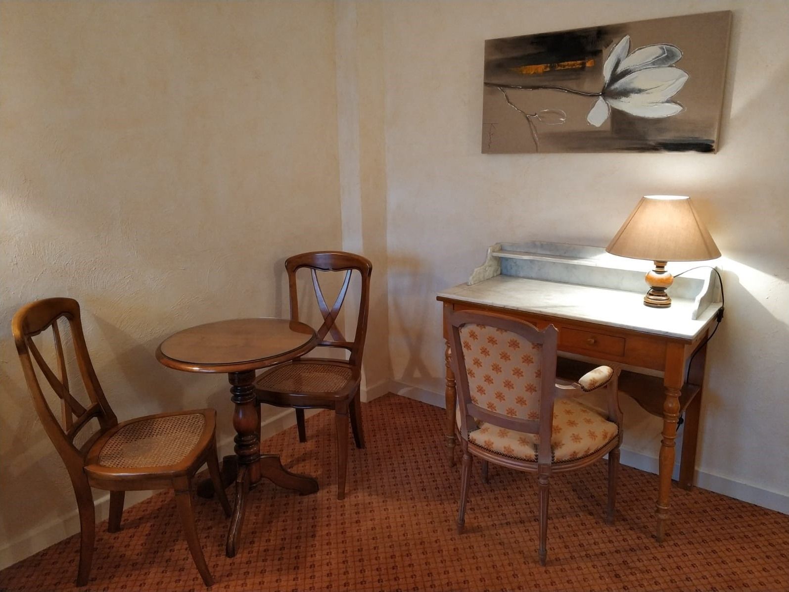 Ein kleines Zimmer mit einem Tisch, zwei Stühlen, einem Schreibtisch mit Lampe und einem Wandbild.