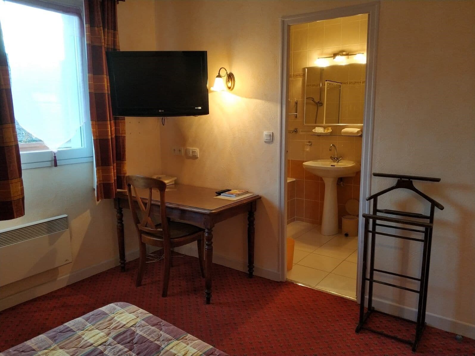 Et hotelværelse med en seng, et skrivebord med stol, et fjernsyn, et badeværelse med håndvask og spejl, samt et tøjstativ.