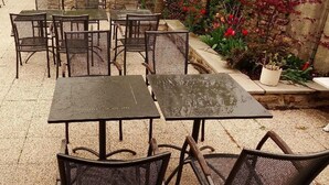 Um pátio externo com mesas e cadeiras de metal, rodeado por um jardim com diversas plantas e flores.