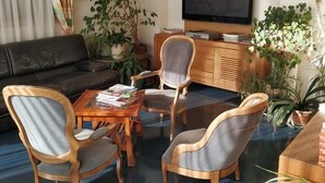 Sala de estar com sofá, cadeiras de madeira, mesa de centro e televisão. Há várias plantas em vasos ao redor.