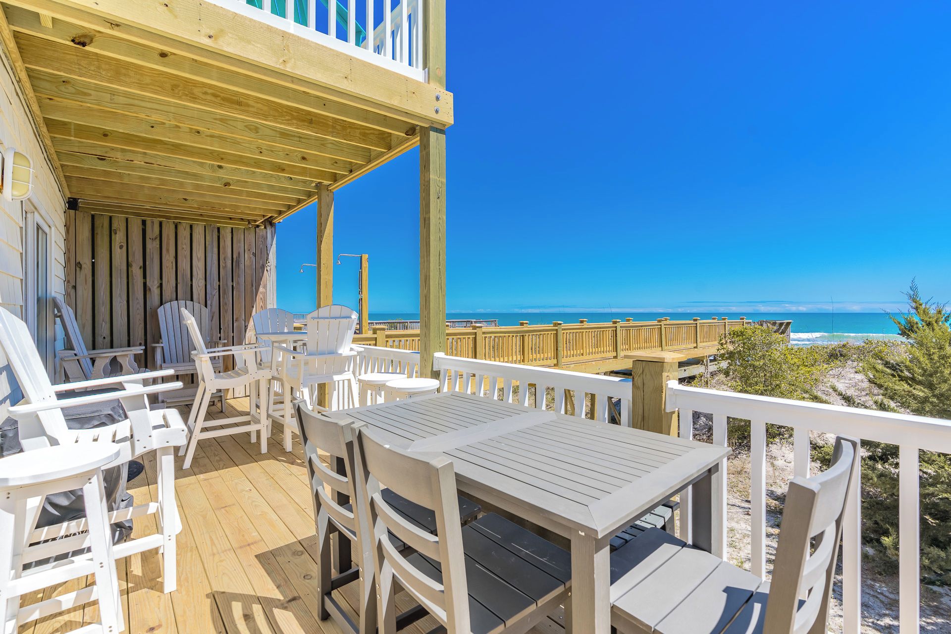 Eine Holzterrasse mit weißen Stühlen und Tischen, mit Blick auf einen Strand.
