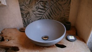 Ein Badezimmer mit einem großen, runden, grauen Waschbecken auf einer Holzplatte, einem kleinen schwarzen Behälter und einer wandmontierten Leuchte.
