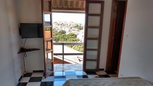 A room with a checkered floor, a bed with a floral bedspread, a TV on the wall, and a balcony with a view of houses and greenery.