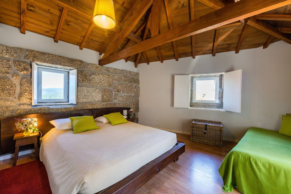 A bedroom with a stone wall, wooden ceiling beams, a large bed with white and green bedding, two windows, and a small bedside table with a lamp.